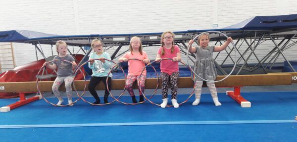 Five children standing inside hula hoops on a blue gym floor, holding ropes in front of a trampoline.