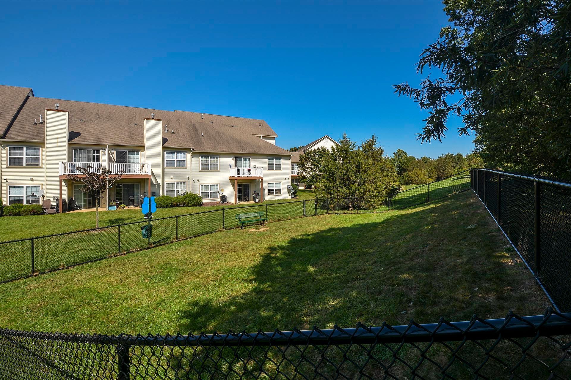 Exterior view of beige apartment buildings with balconies, a grassy yard, and a chain-link fence under a blue sky.