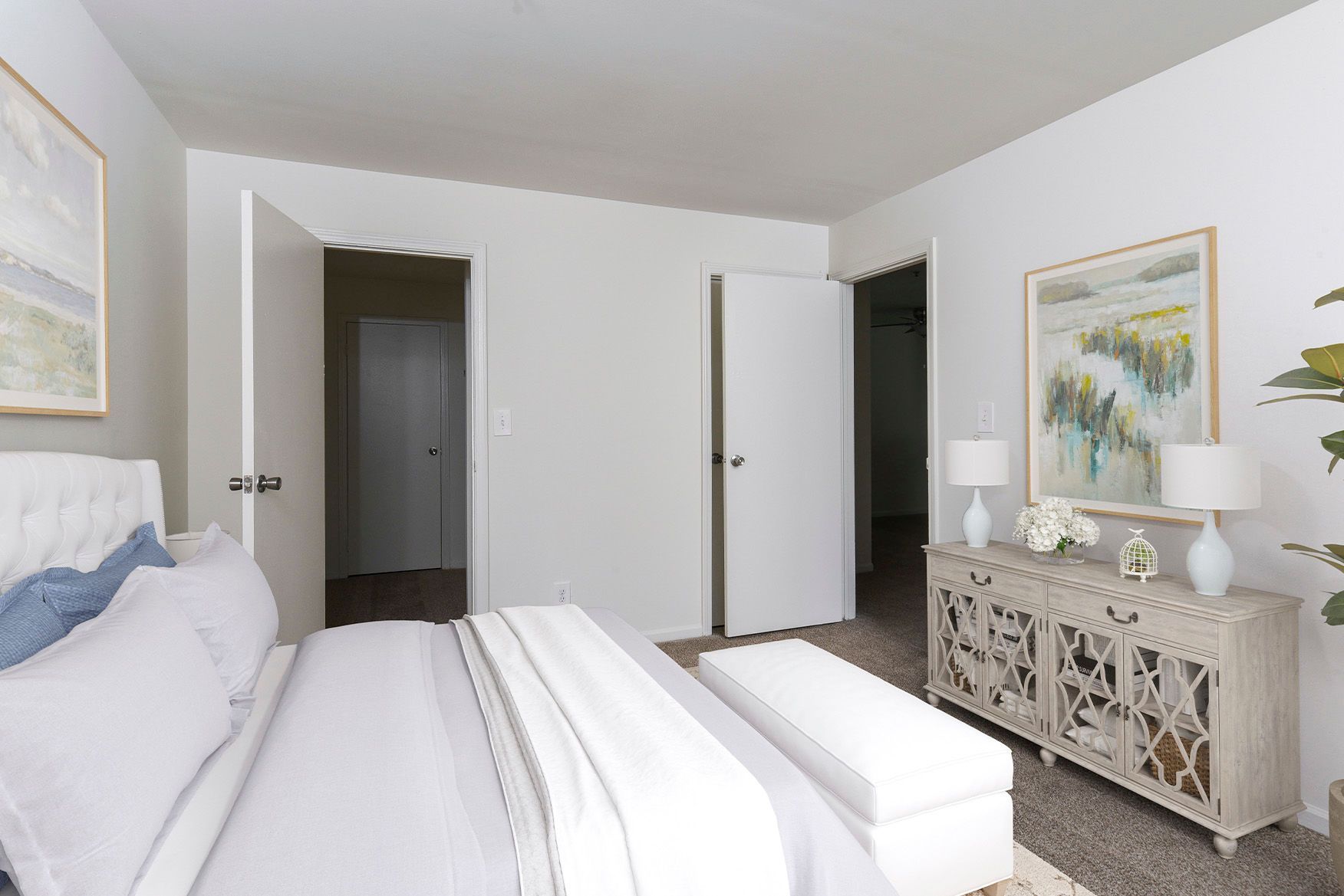 Modern apartment bedroom with white upholstered bed, neutral decor, and two open doors.
