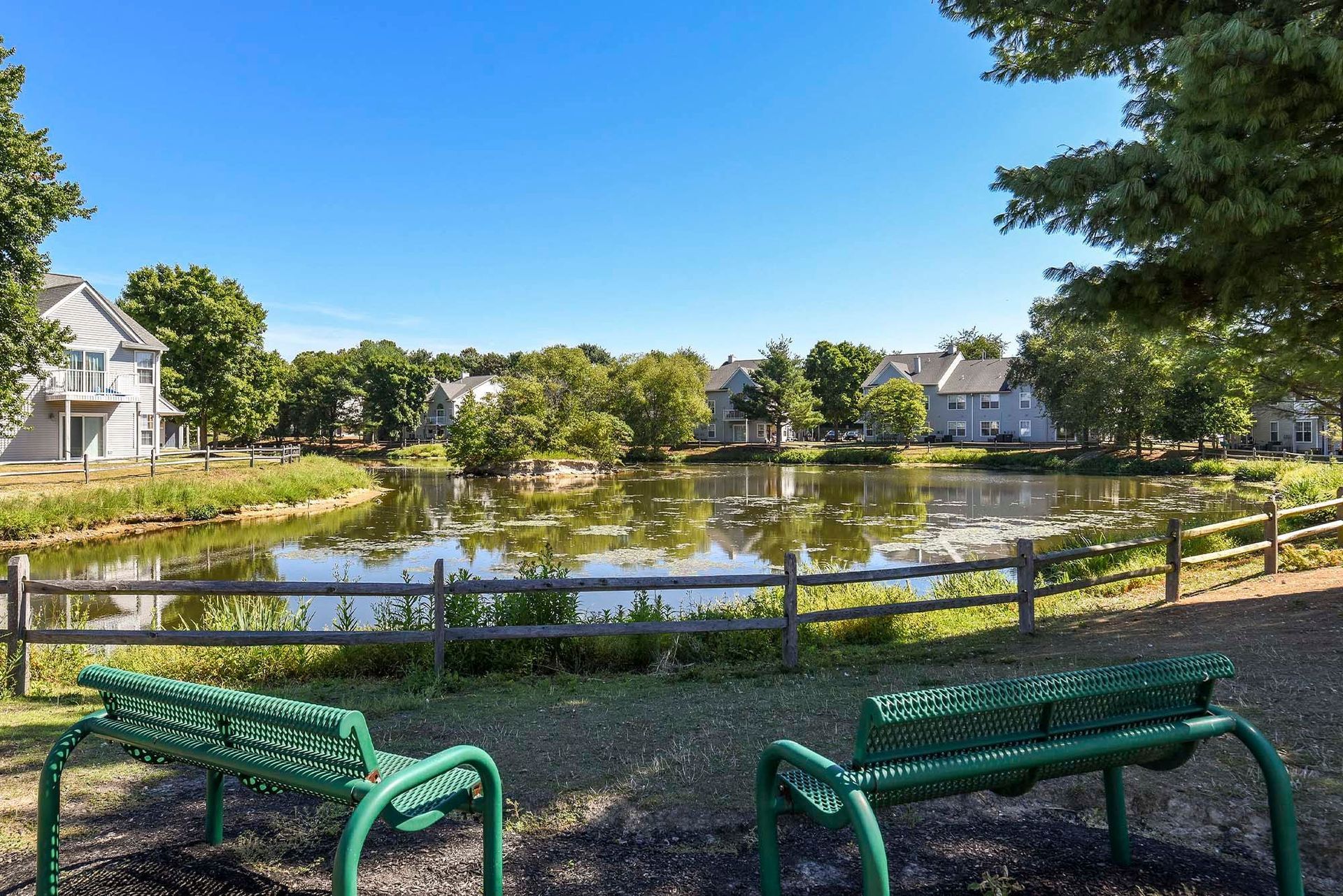 Outdoor pond with green benches and trees, surrounded by apartment buildings.