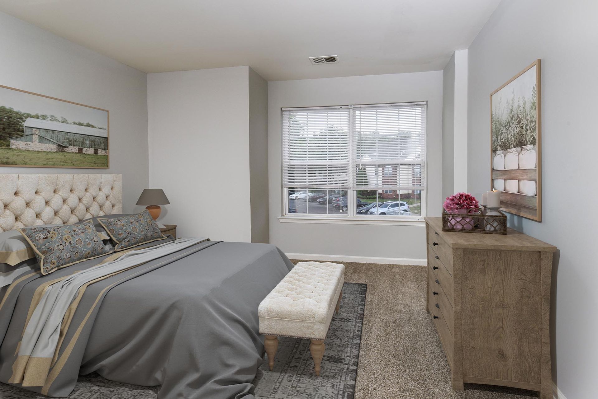 Bedroom with tufted beige headboard, gray bedding, bench at foot, and wooden dresser by window.