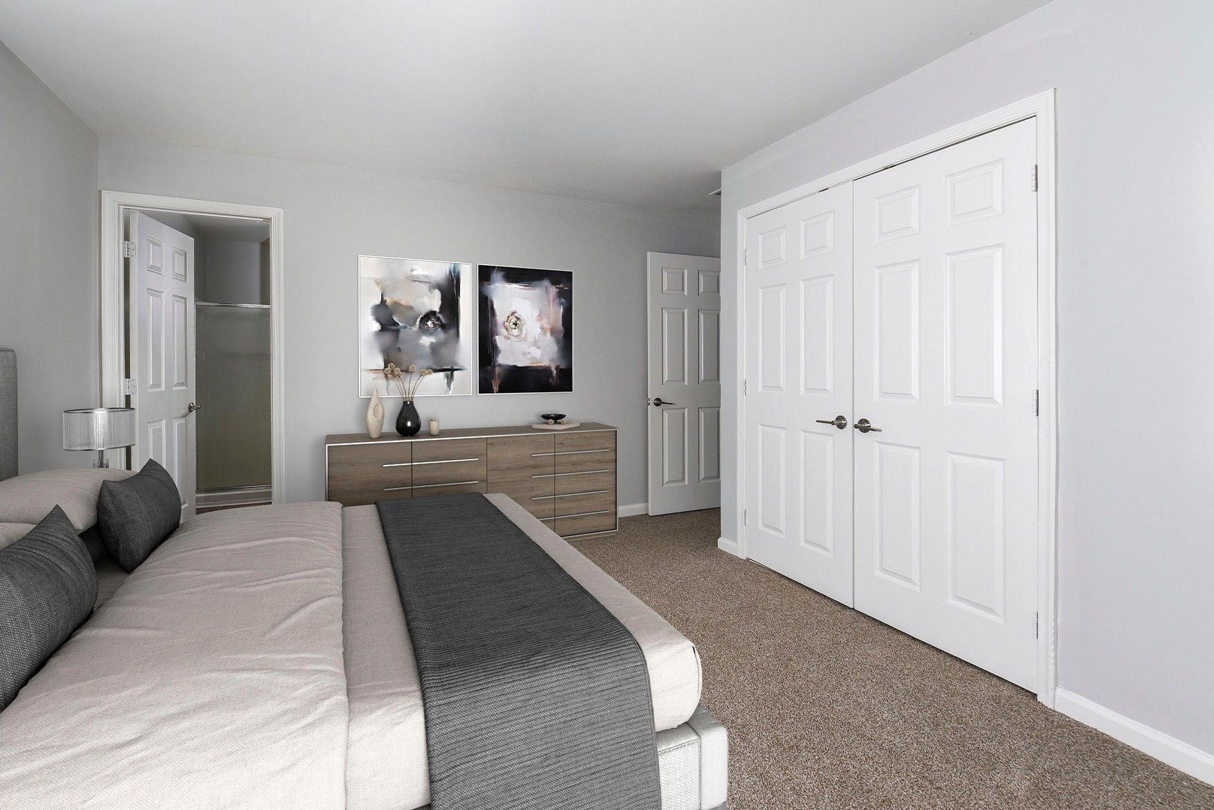 Bedroom in apartment with a large bed, neutral walls, a wooden dresser, and abstract wall art.