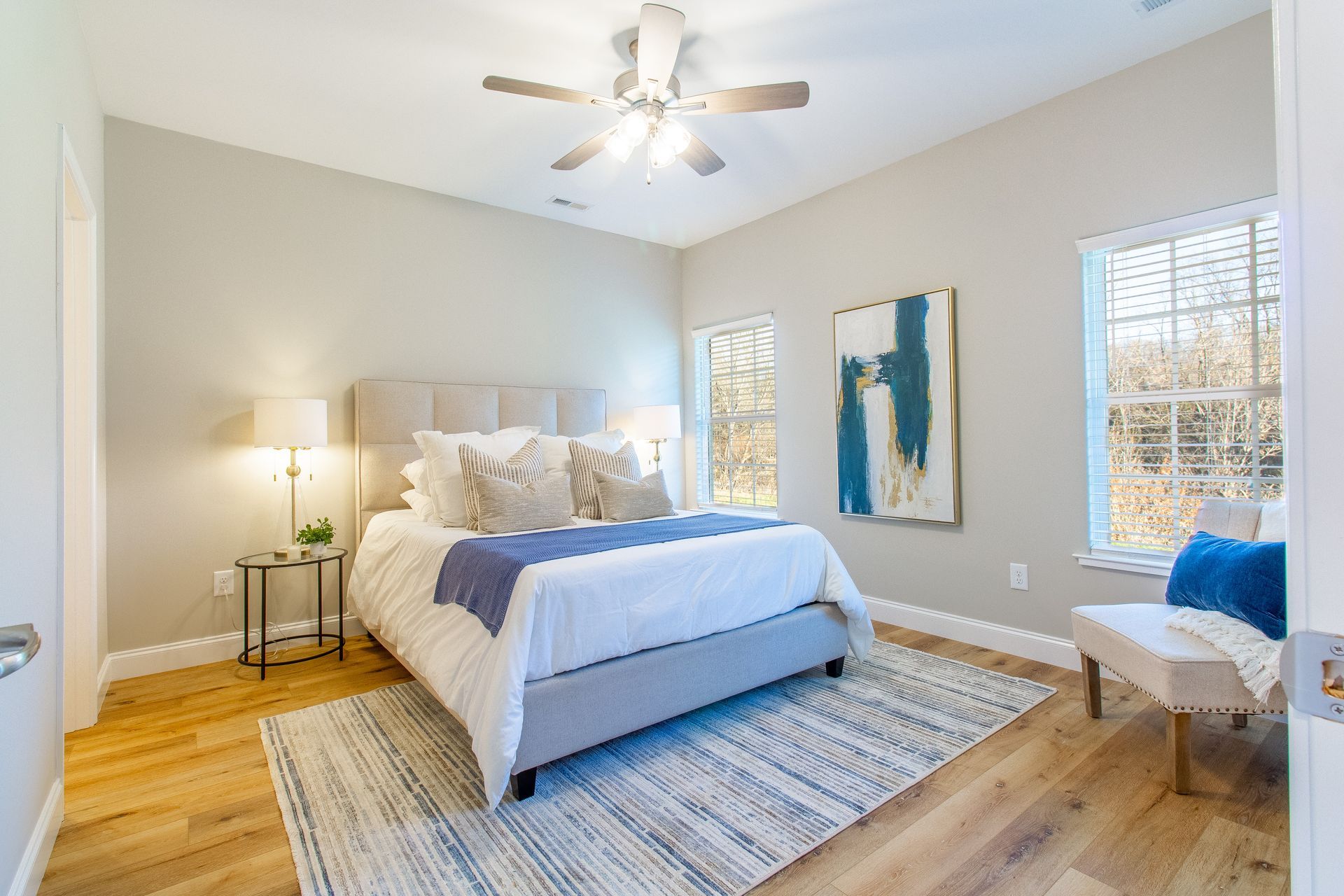 A bedroom with a king size bed , a rug , a ceiling fan and two windows.
