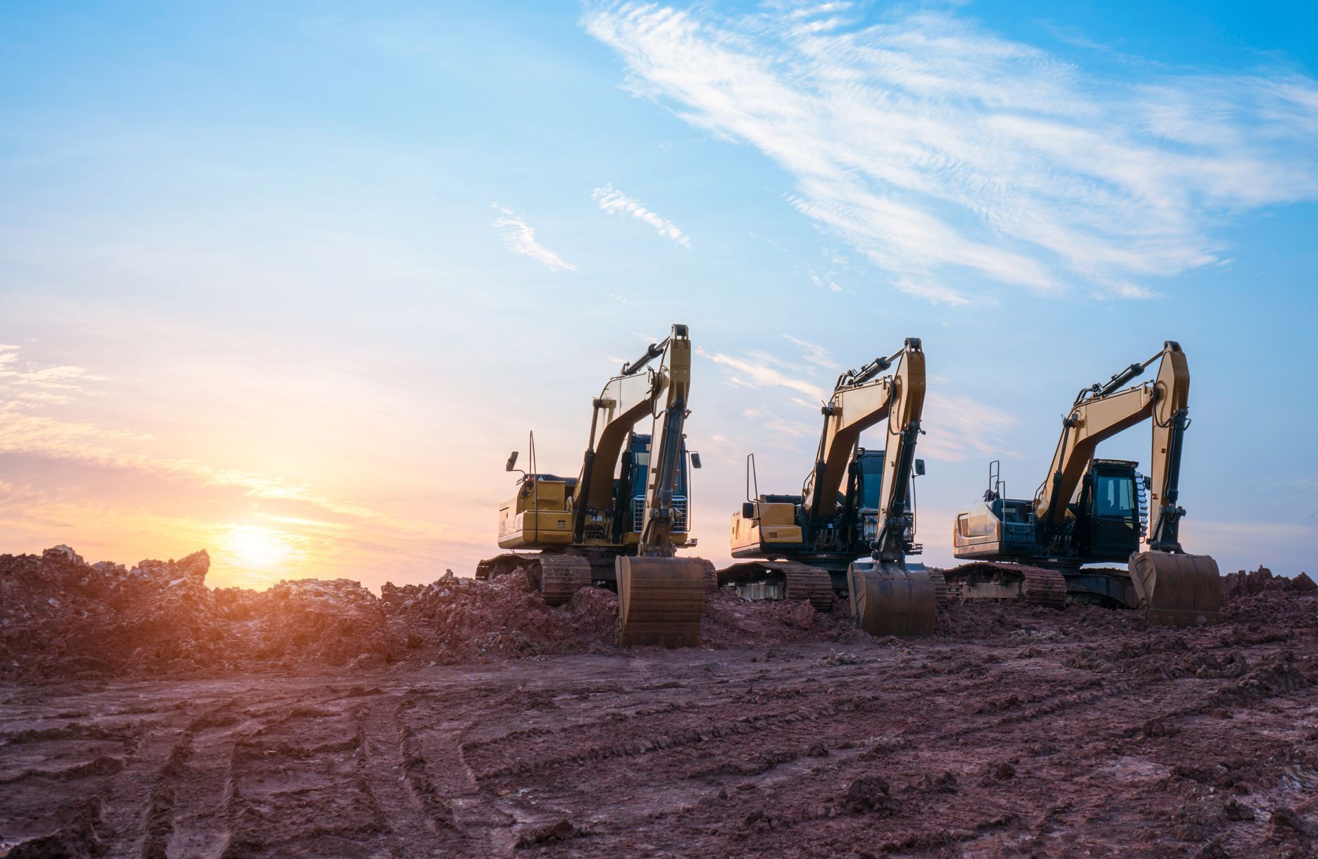 Three yellow excavators on a muddy construction site at sunrise under a blue sky.