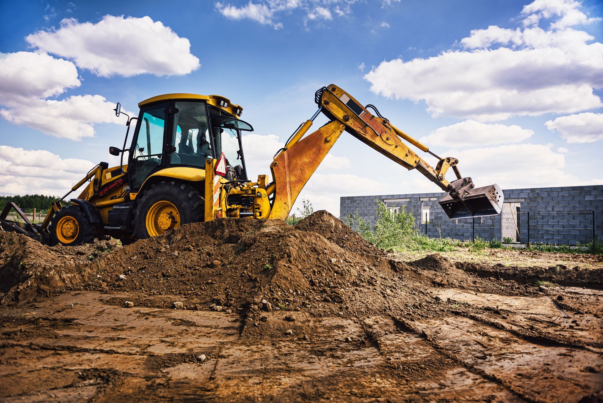 Yellow backhoe digging in dirt under a blue sky with clouds.