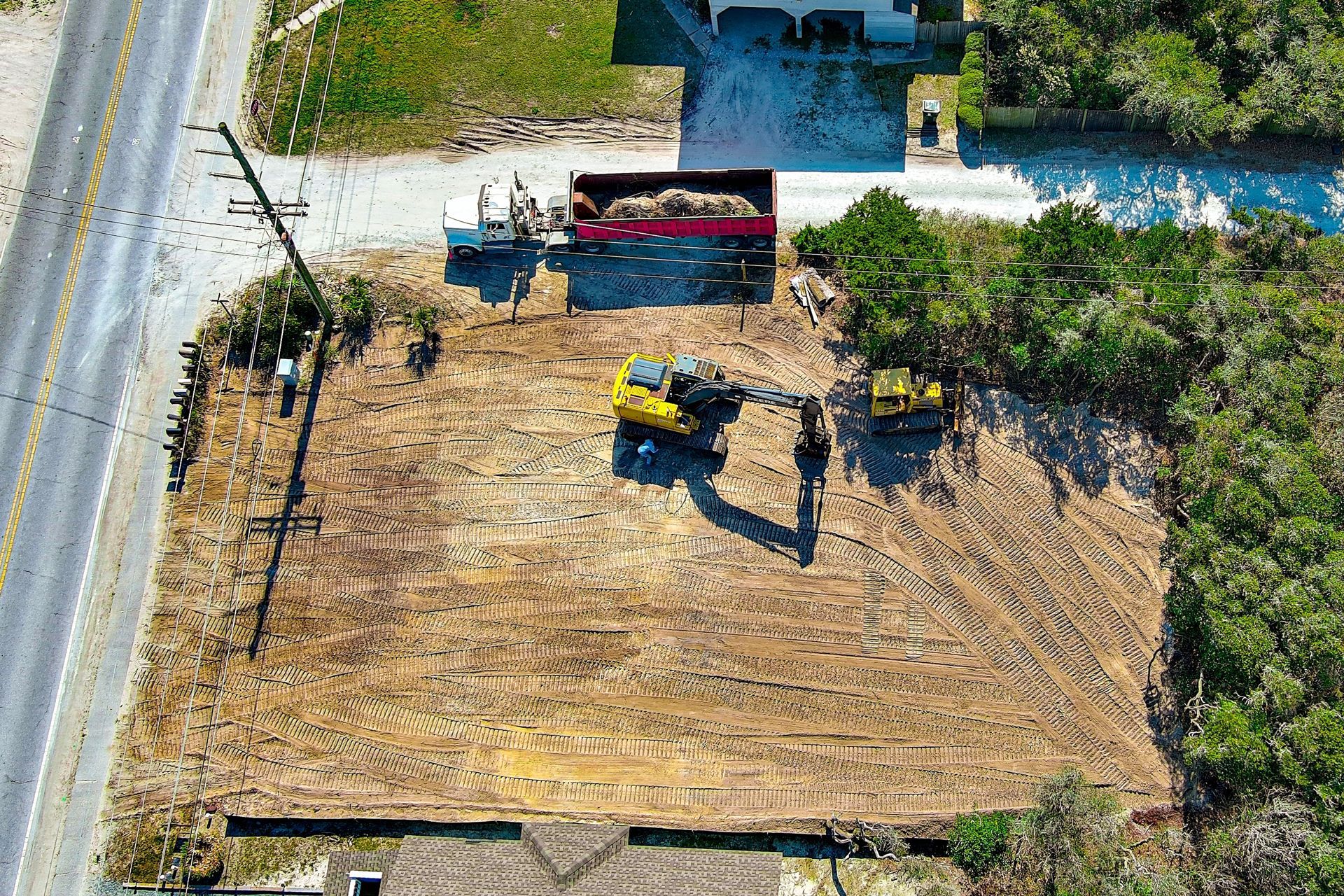 Construction site with excavator and truck removing soil; road, trees, and power lines visible.