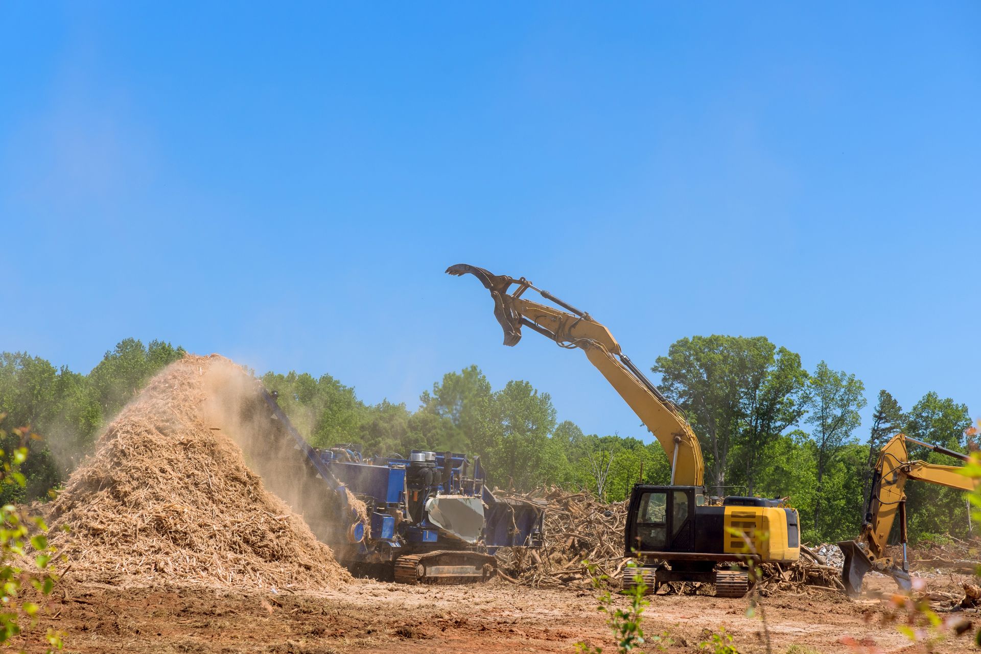 Excavator and wood chipper clearing trees, creating wood chips under a bright blue sky.