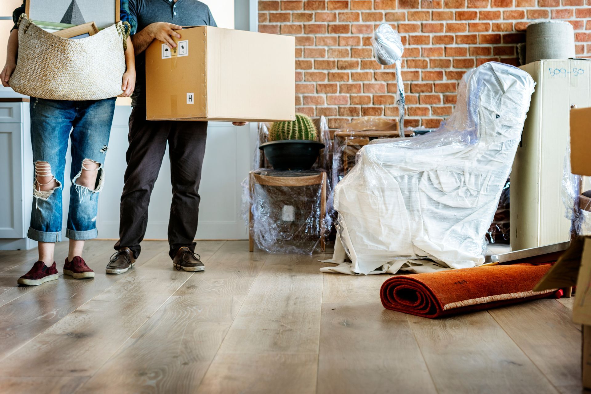  Two people carrying moving boxes in a room with packed furniture, a rolled-up rug, and a brick wall