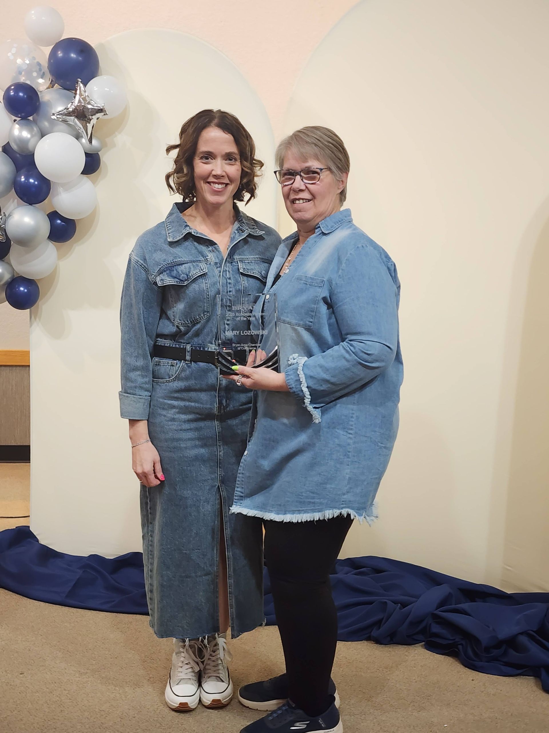 Two women in denim shirts smile while standing together in front of a blue and white balloon decoration.