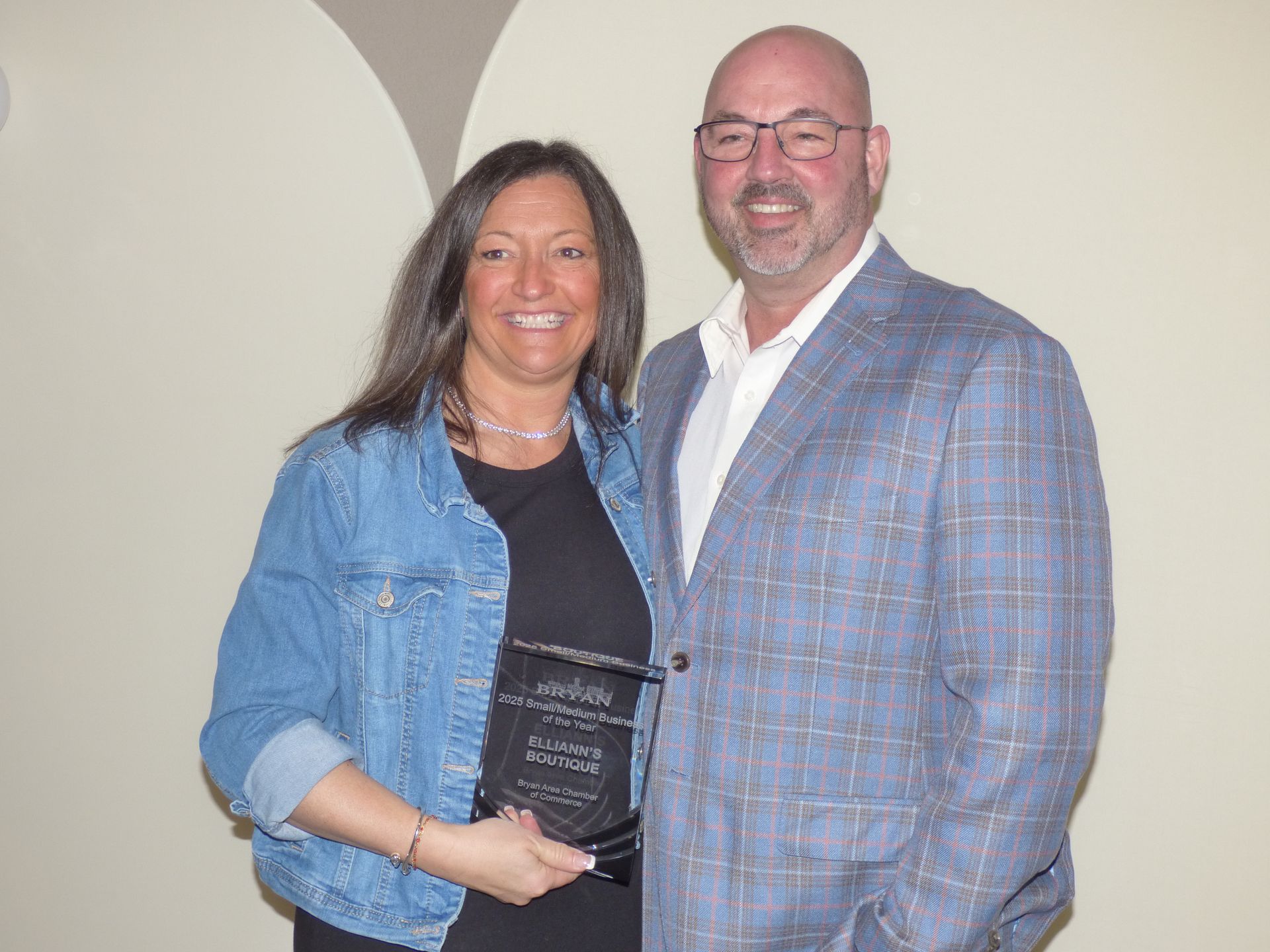 A smiling woman holding a dark award plaque stands next to a smiling man in a blue plaid blazer against a plain wall.