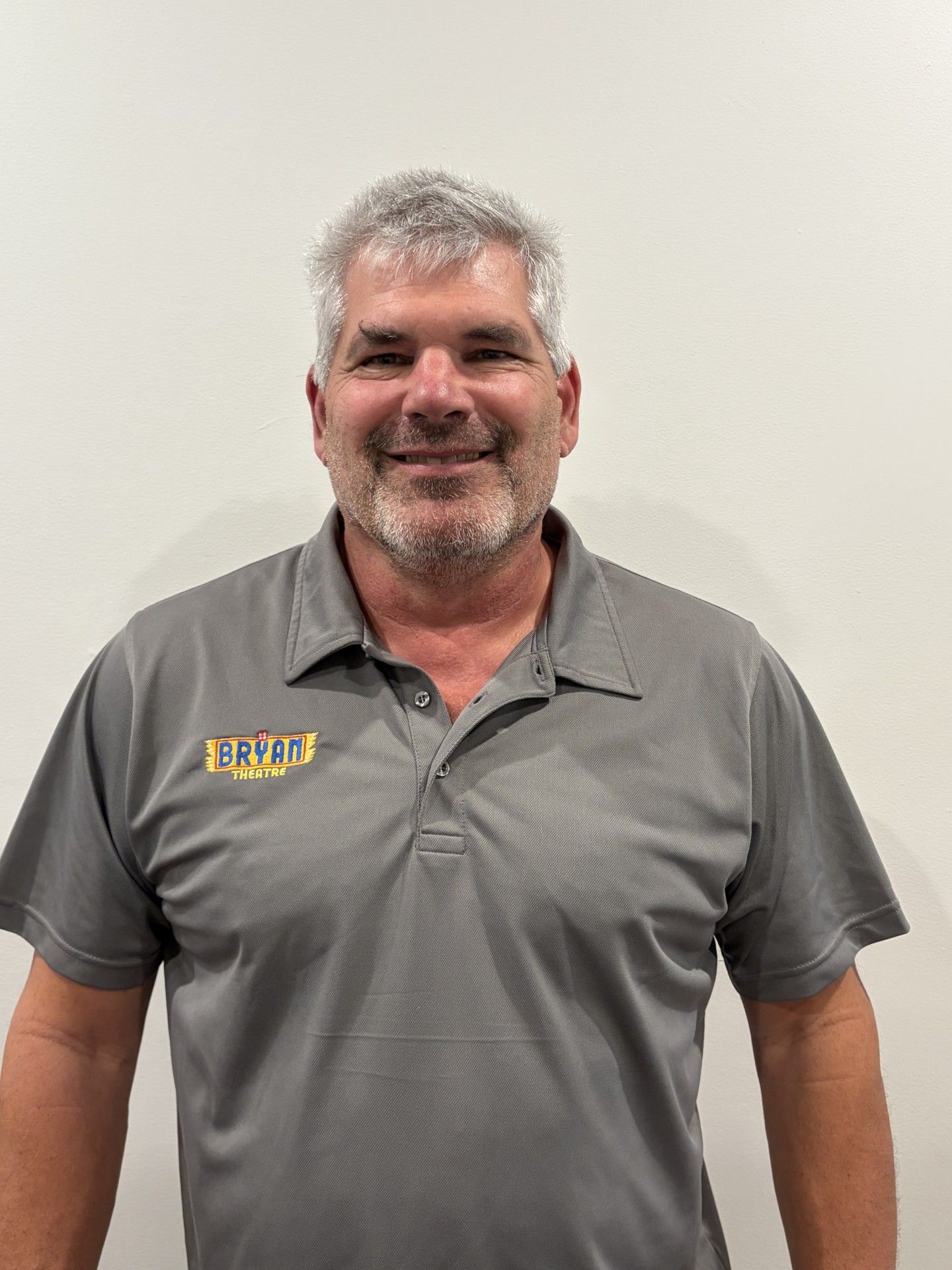 A smiling person with gray hair and facial hair, wearing a gray polo shirt with a yellow logo, stands against a white wall.