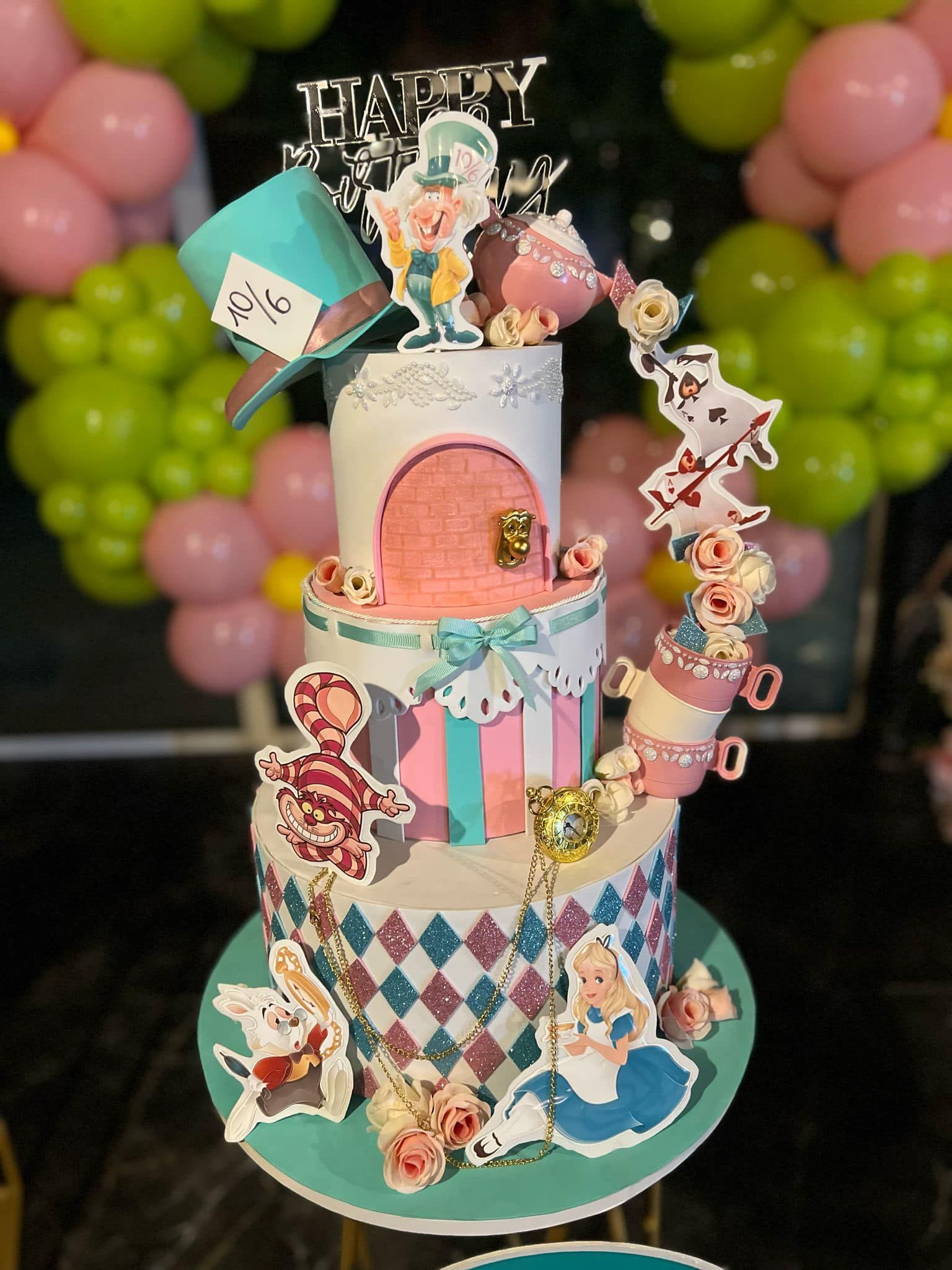Torta a tre piani ispirata ad Alice nel Paese delle Meraviglie, con decorazioni e personaggi; rosa, blu e bianco; palloncini sullo sfondo.