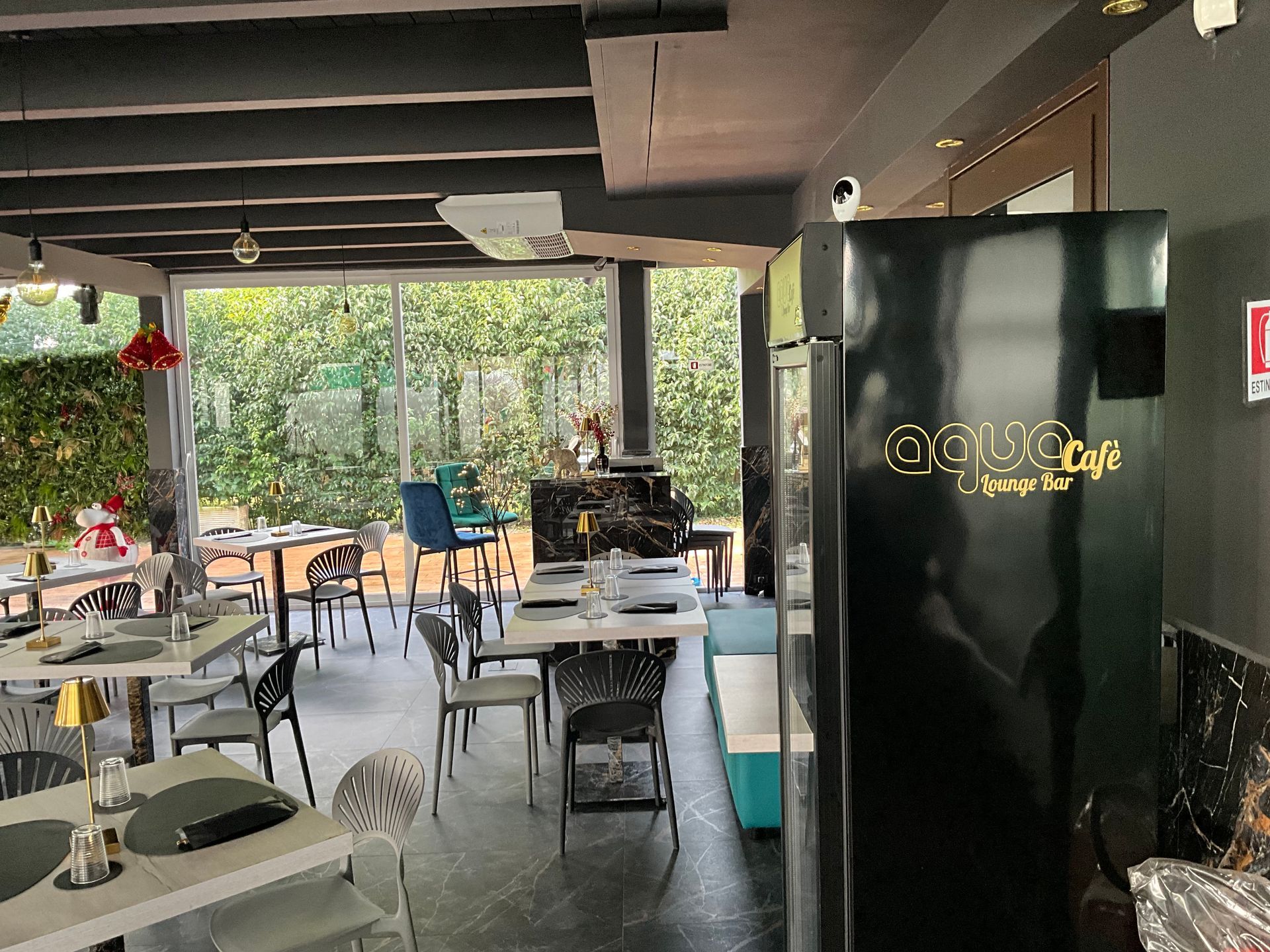Interno dell'Aqua Café con tavoli, sedie e un grande frigorifero. Le vetrate offrono una vista sul verde.