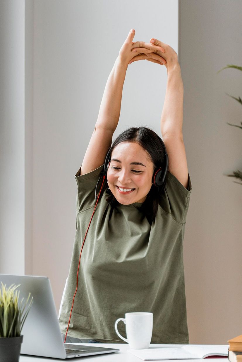 A woman is stretching her arms while sitting at a desk in front of a laptop computer.