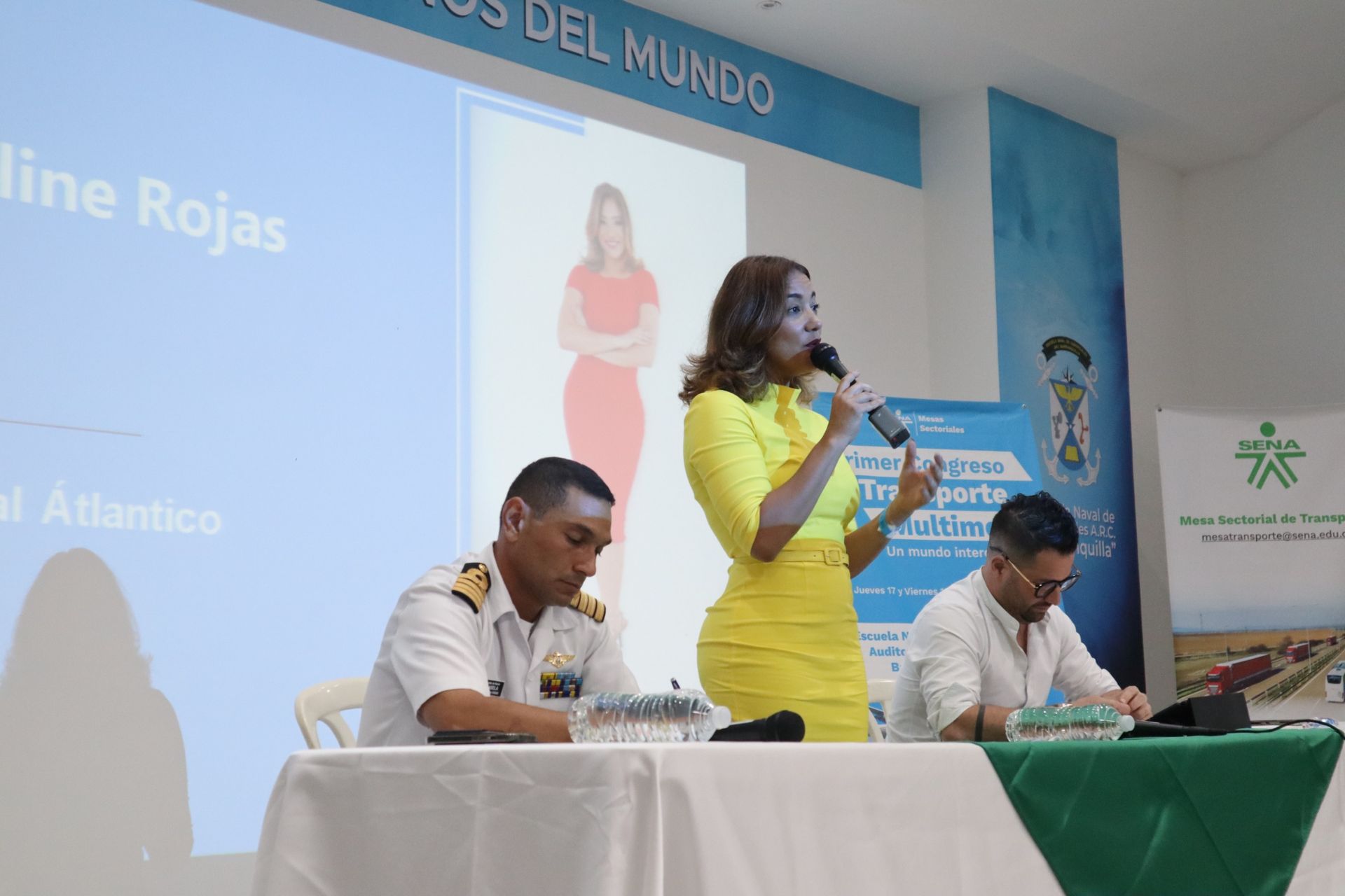 Una mujer con un vestido amarillo habla a través de un micrófono mientras dos hombres están sentados en una mesa.