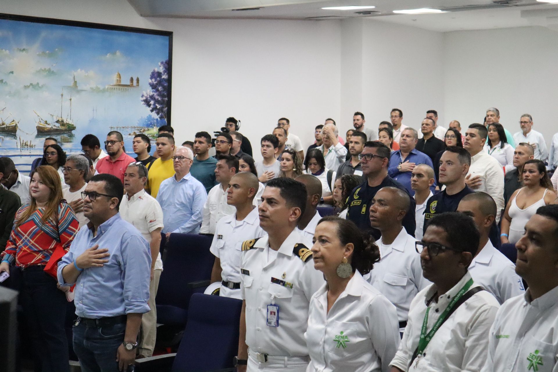 Un gran grupo de personas se encuentran en una habitación con las manos en el corazón.