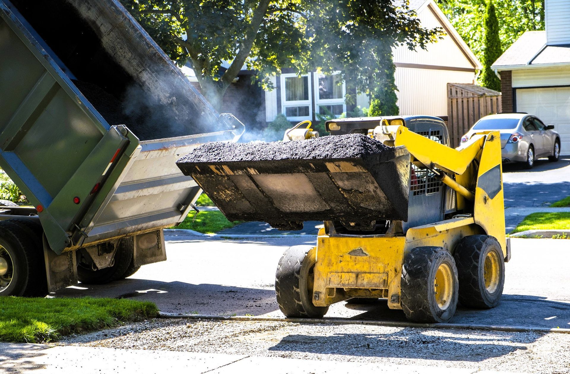 A skid-steer loader carrying gravel from a dump truck for road paving.