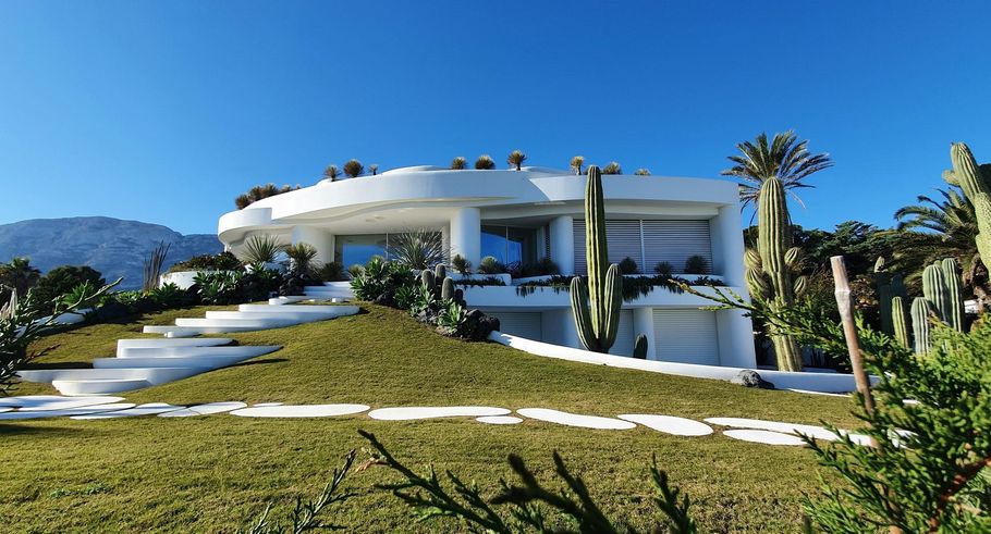 Modern white hillside villa with curved architecture, palm trees, and a landscaped lawn under a clear blue sky