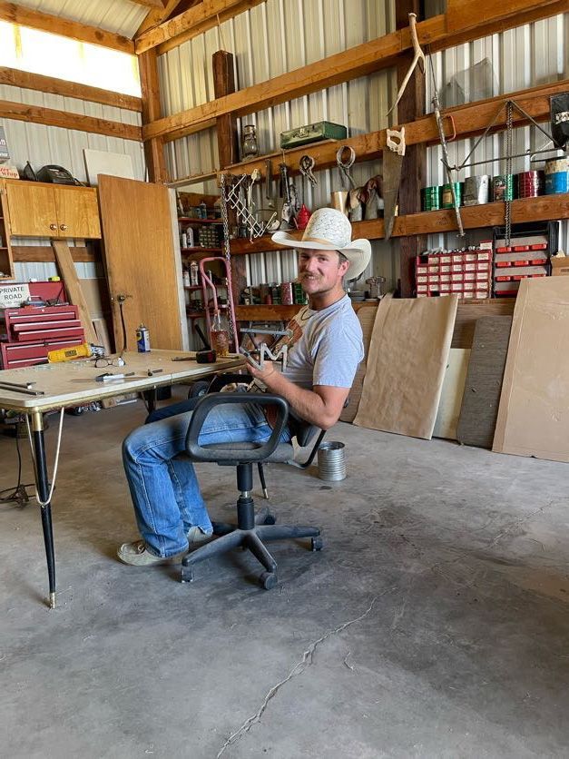 Man in a cowboy hat smiles while seated at a table in a workshop, surrounded by tools.