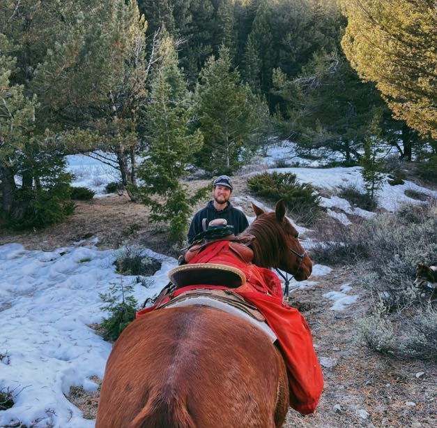 Man riding a brown horse on a snowy trail in a forest, smiling. Horse is wearing a red blanket.