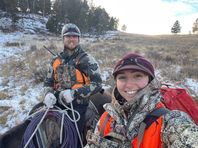 Two hunters smiling on horseback in a snowy field. One holds rope. Both wear camo and orange vests.