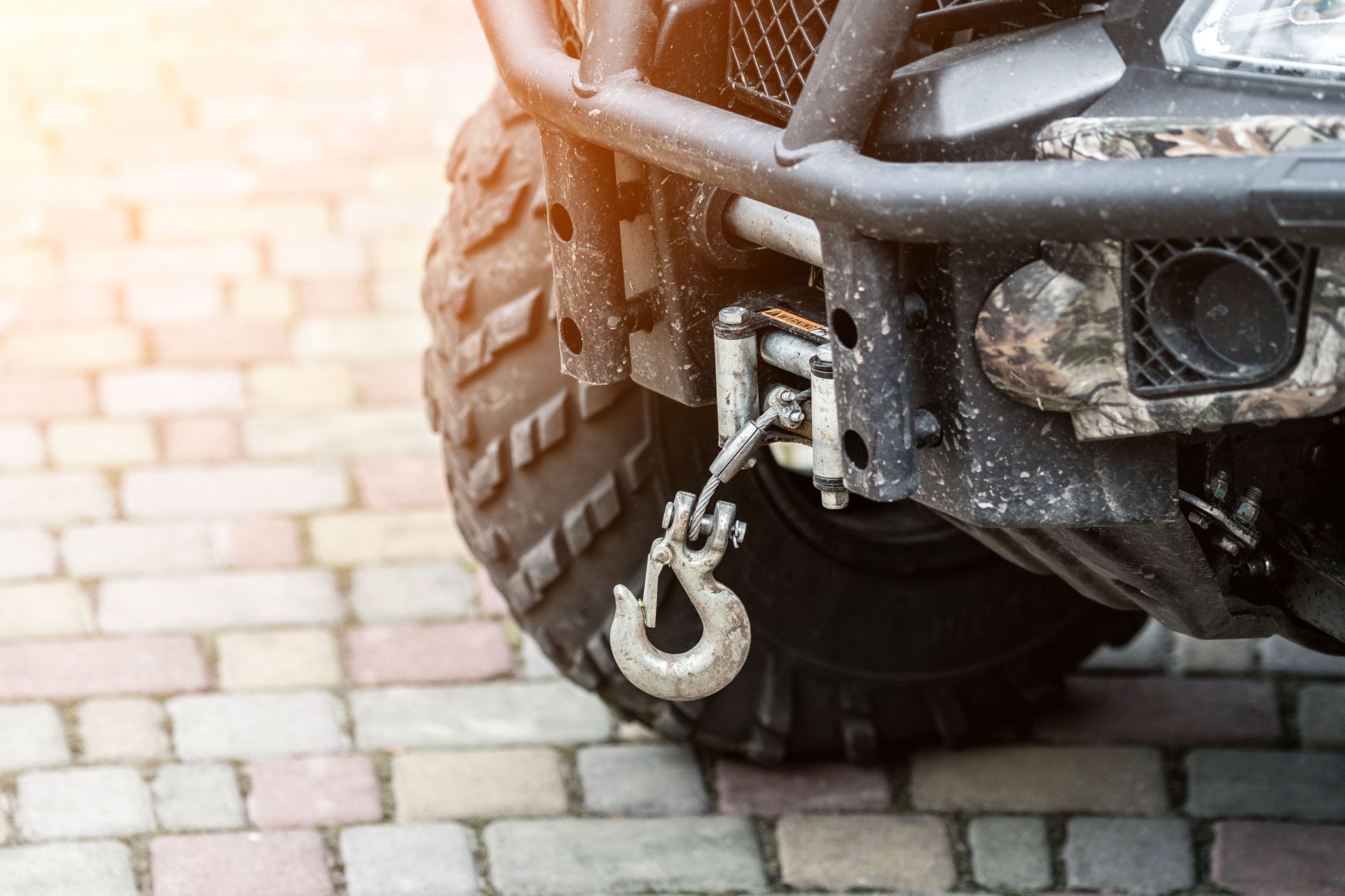 Close-up of an ATV's front bumper, winch, and tire on a brick surface.