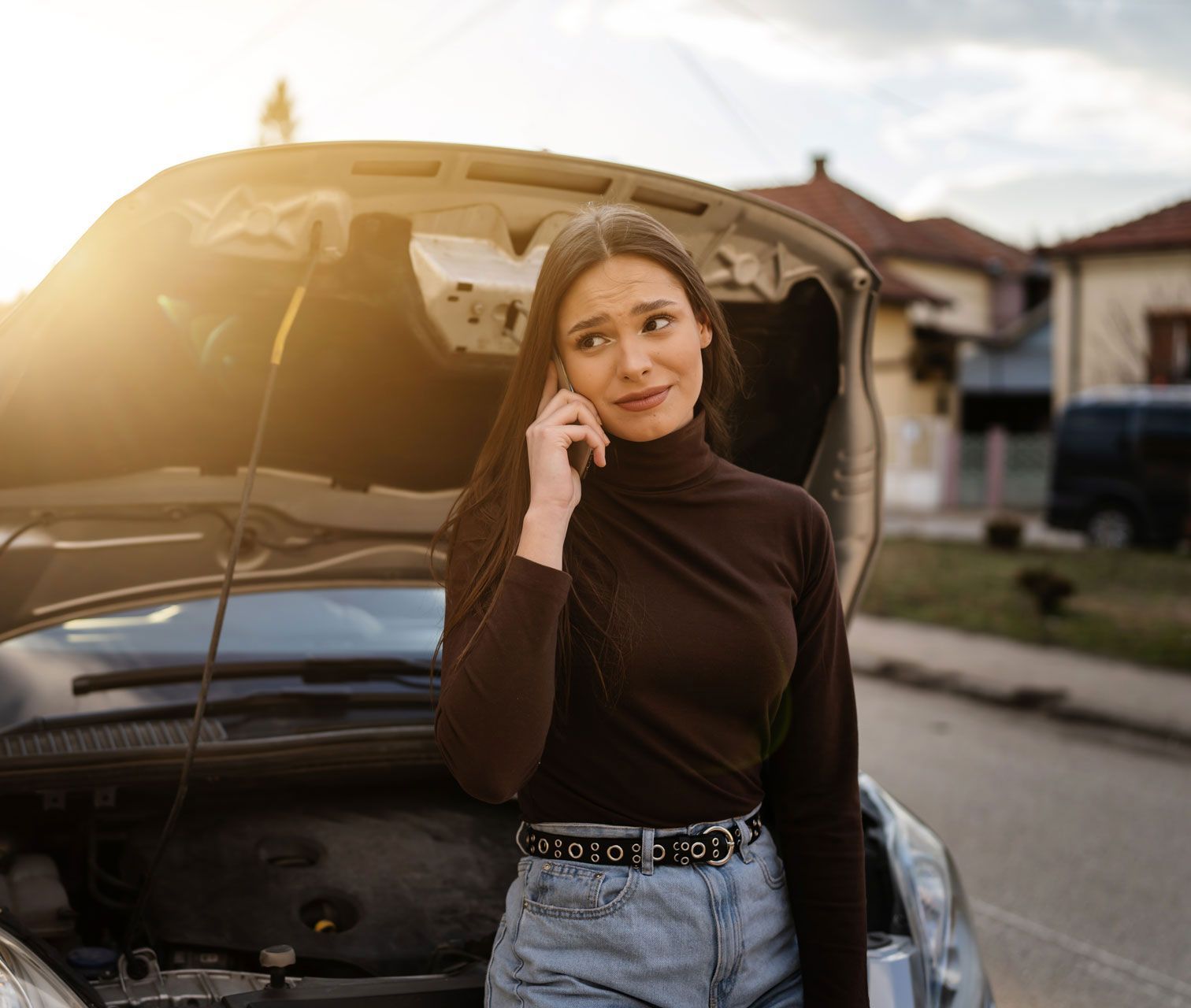 Woman with open car hood talking on phone, looking concerned on a residential street.