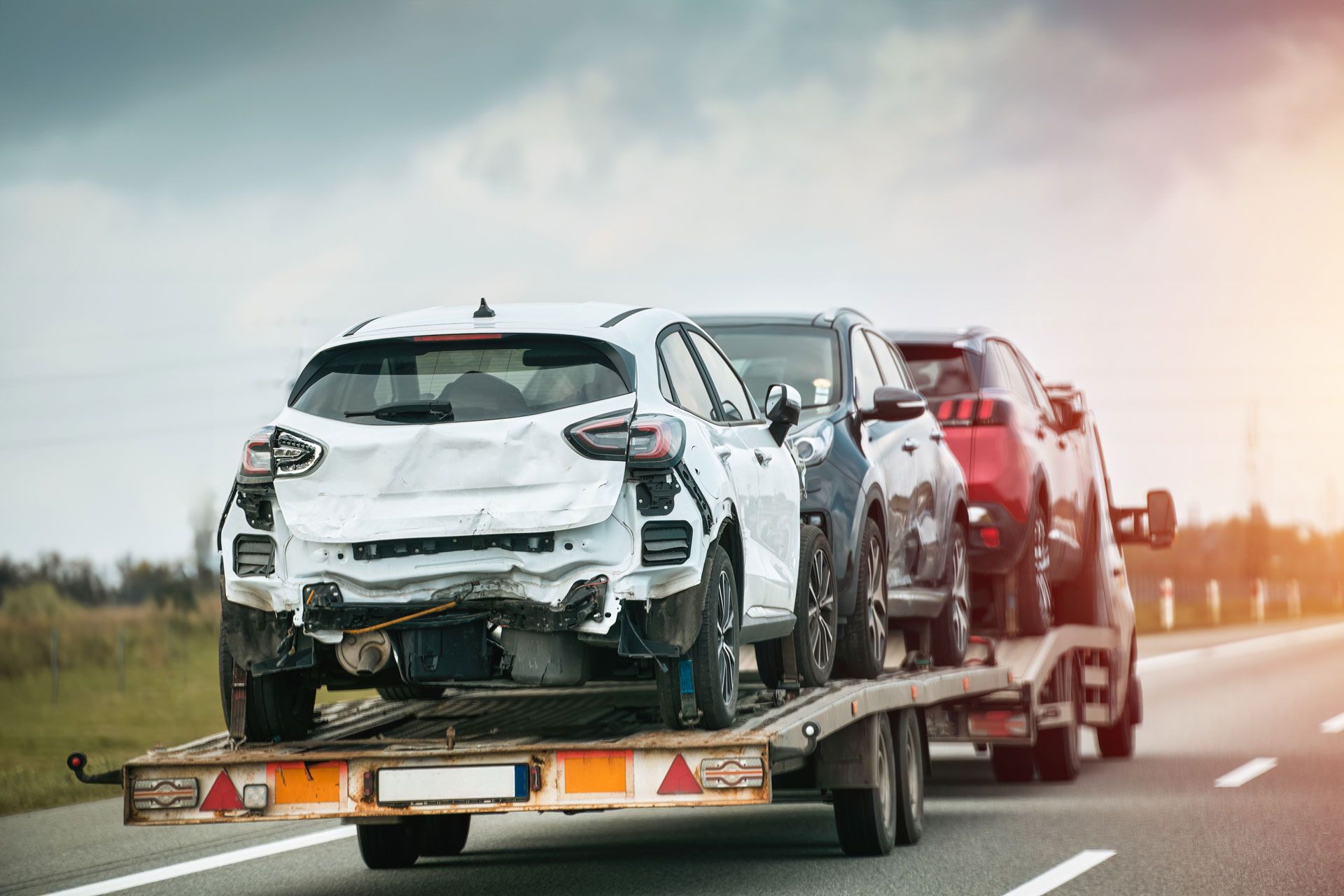 Cars on a flatbed trailer traveling on a highway, one car severely damaged.
