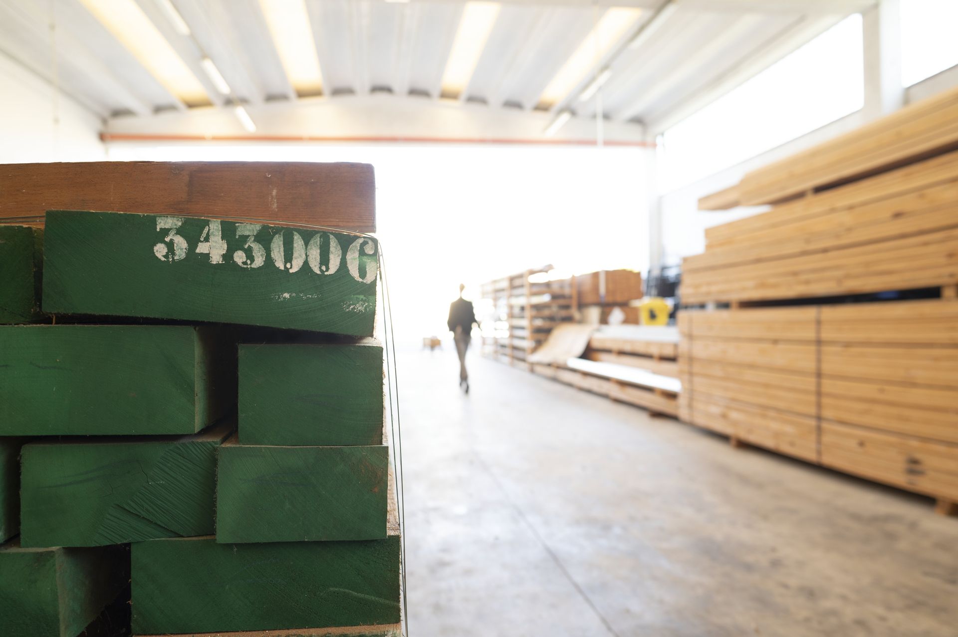 Catasta di legname verde con numero bianco, interno del magazzino, persona che cammina verso l'uscita.