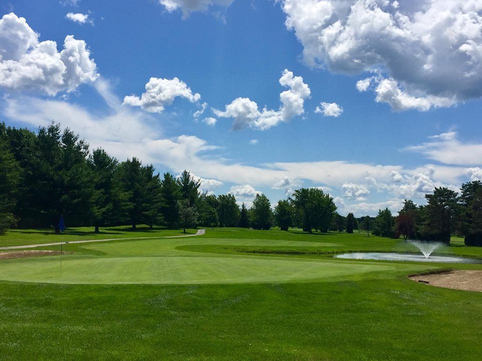 Green golf course under a blue sky with white clouds, trees, and a water fountain.