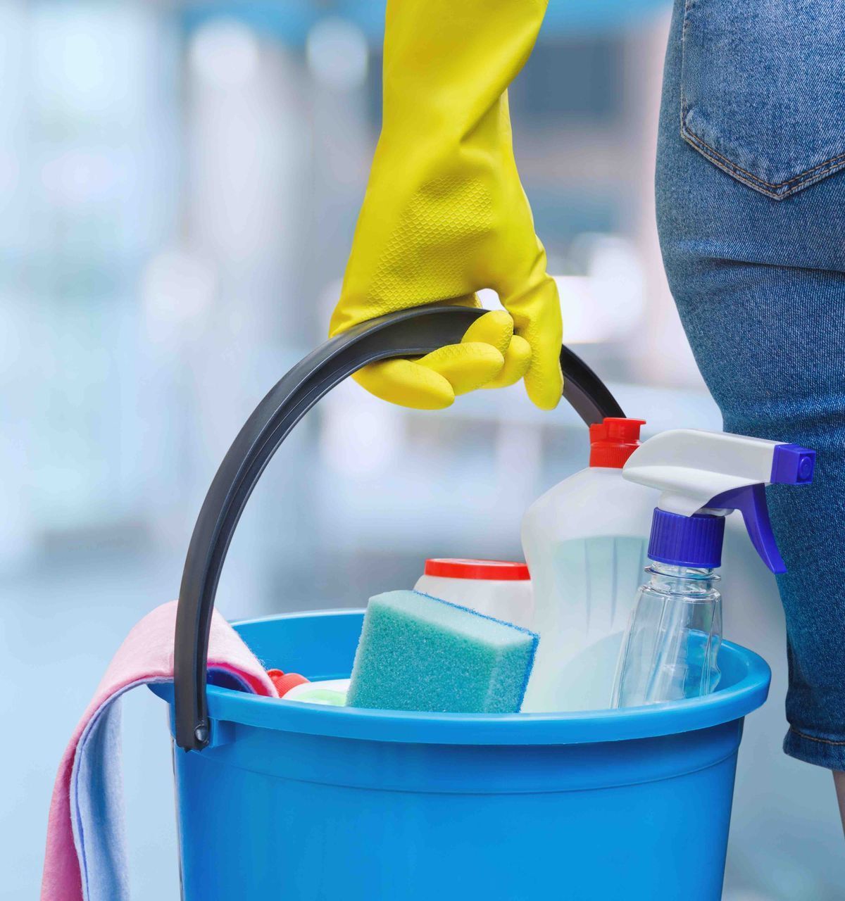 A person wearing yellow gloves is holding a blue bucket filled with cleaning supplies.