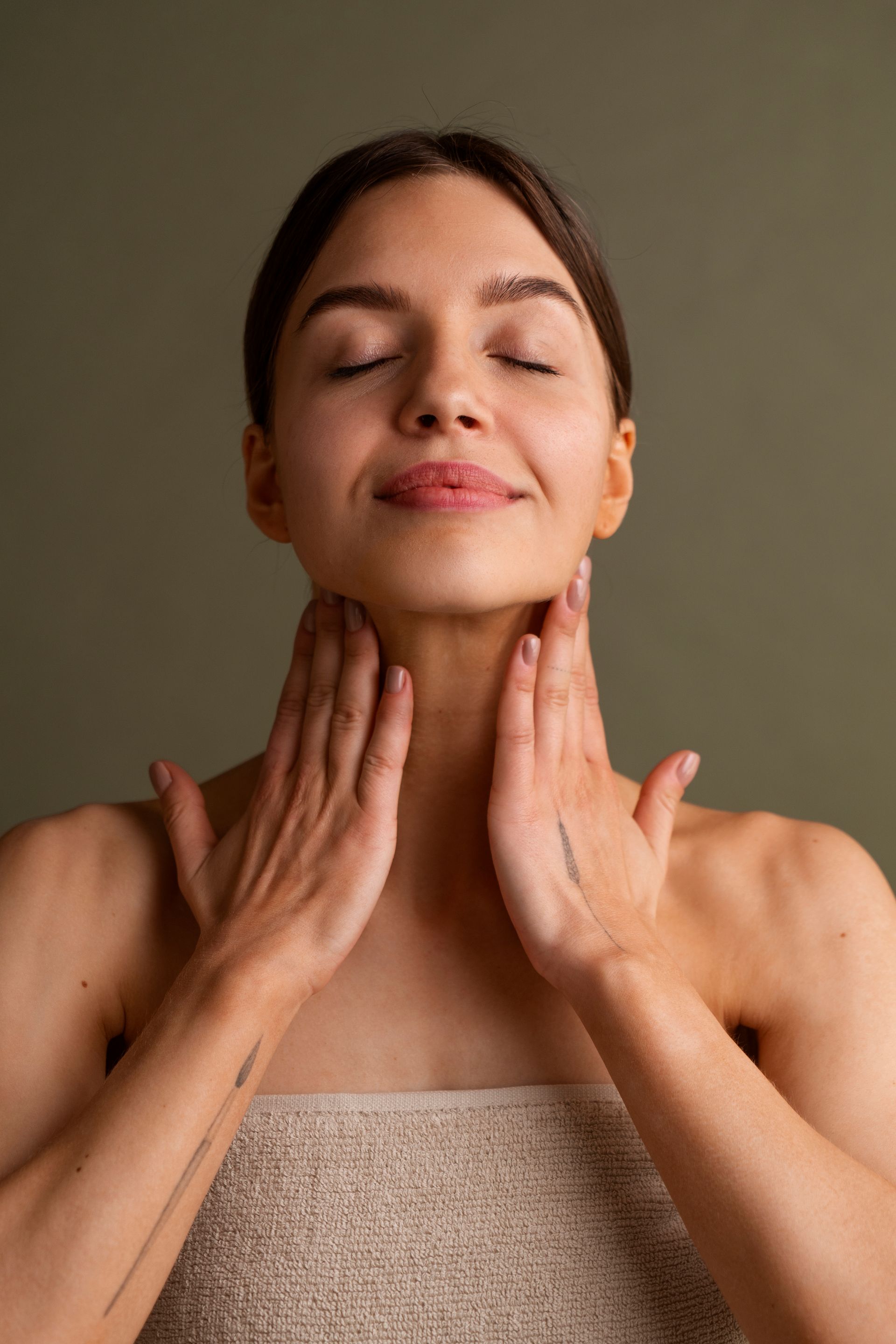 Mujer con ojos cerrados, manos en el cuello, tocando la piel, expresión suave, toalla.