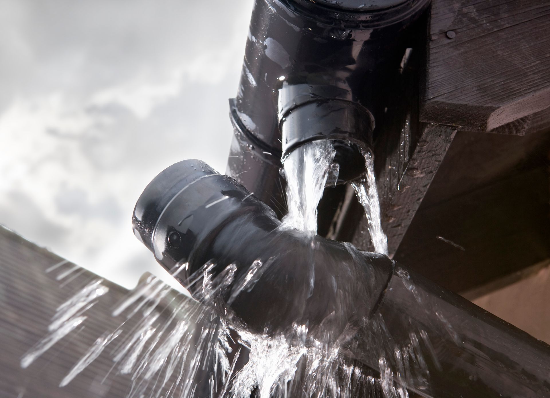 Water gushes from a black gutter downspout onto a wooden surface under a cloudy sky.