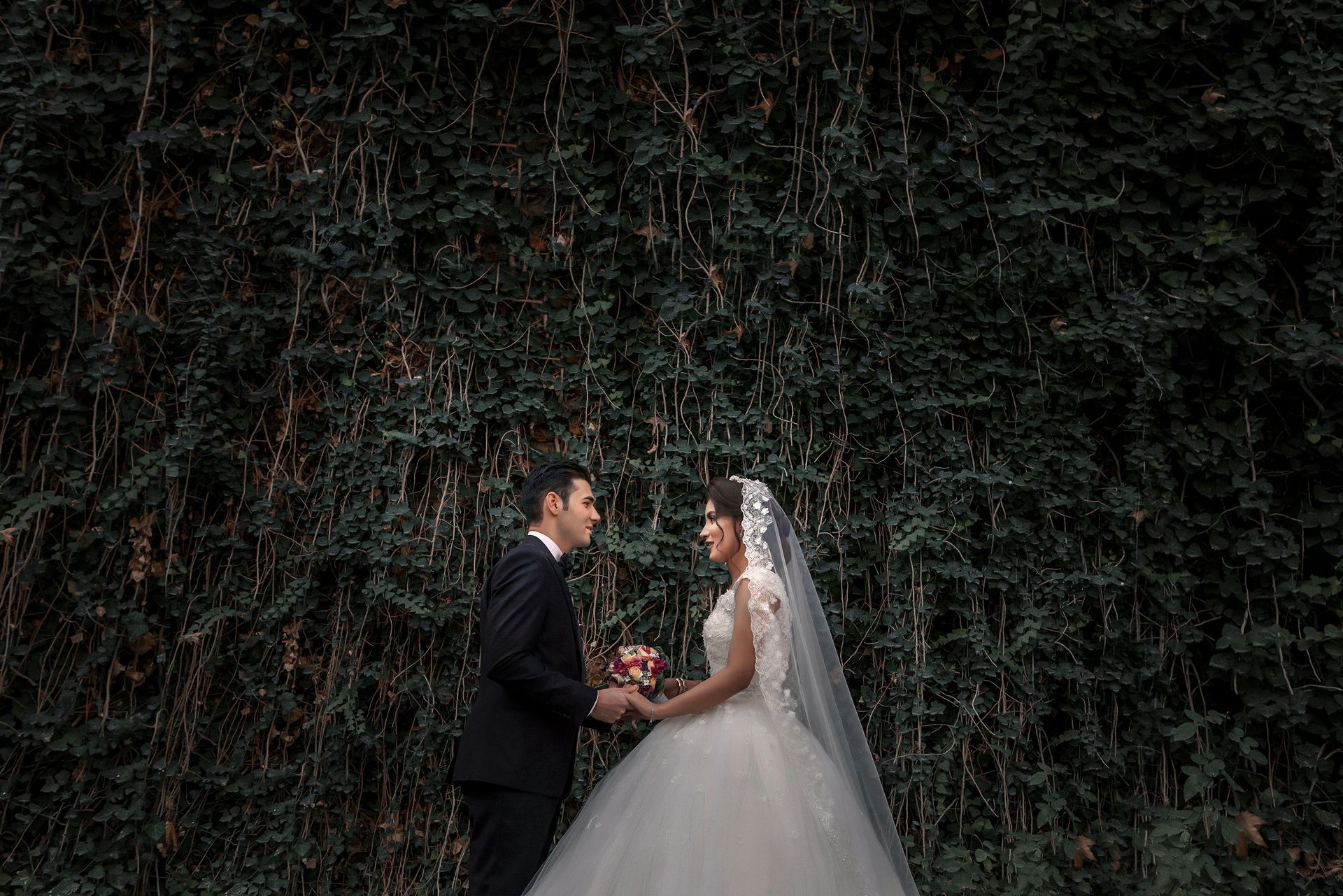 A bride and groom are holding hands in front of a wall of trees.