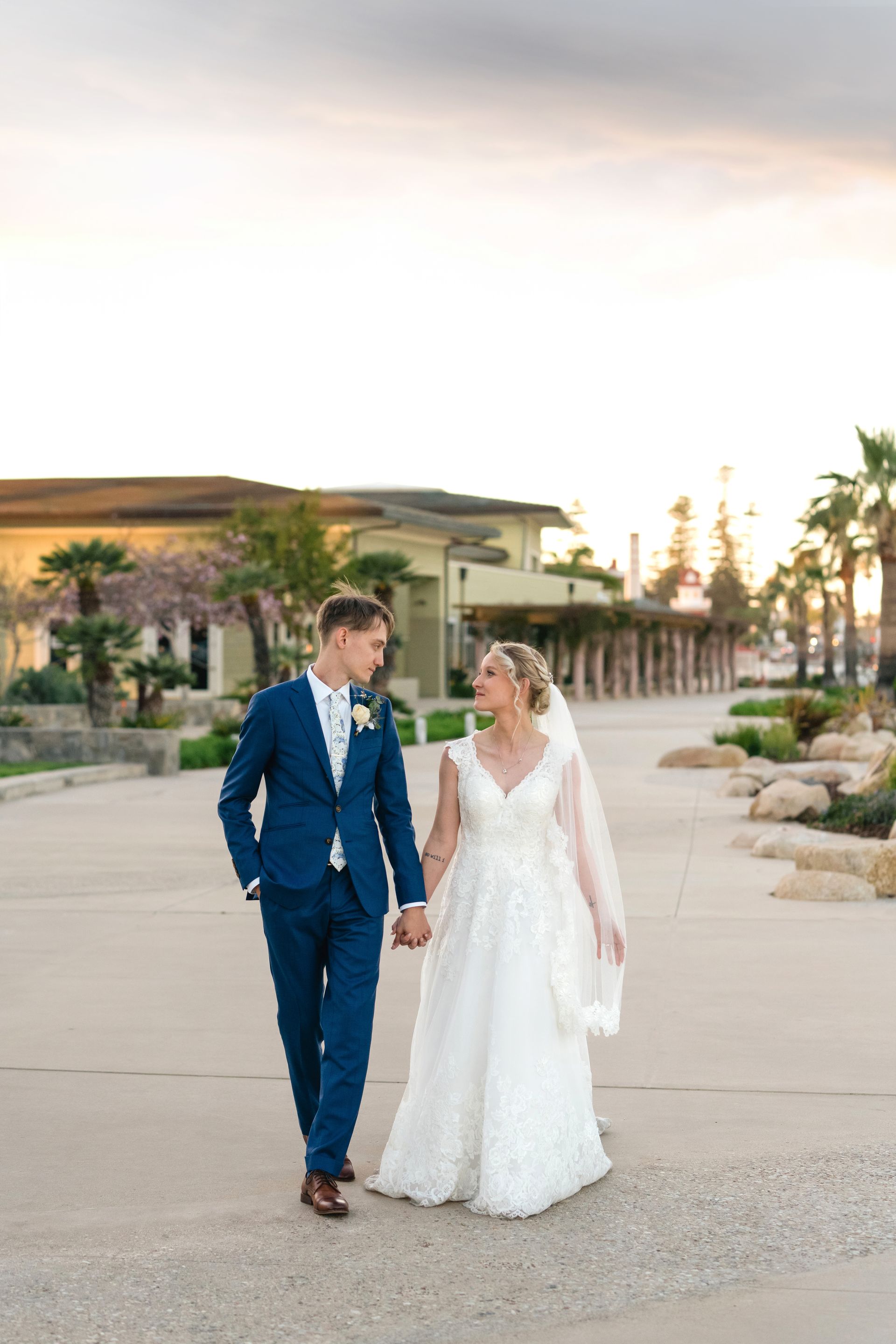 A bride and groom are walking down a sidewalk holding hands.