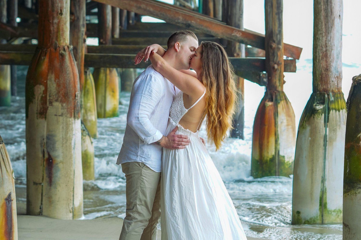 A man and a woman are kissing under a pier on the beach.