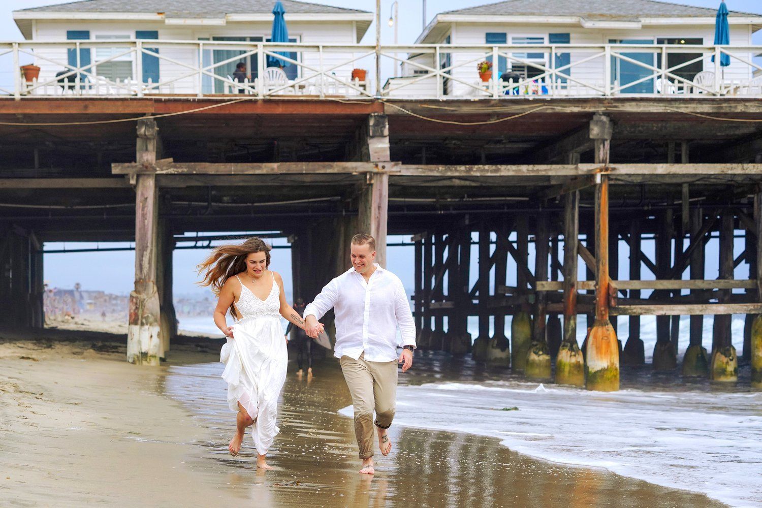 A man and a woman are running on the beach under a pier.