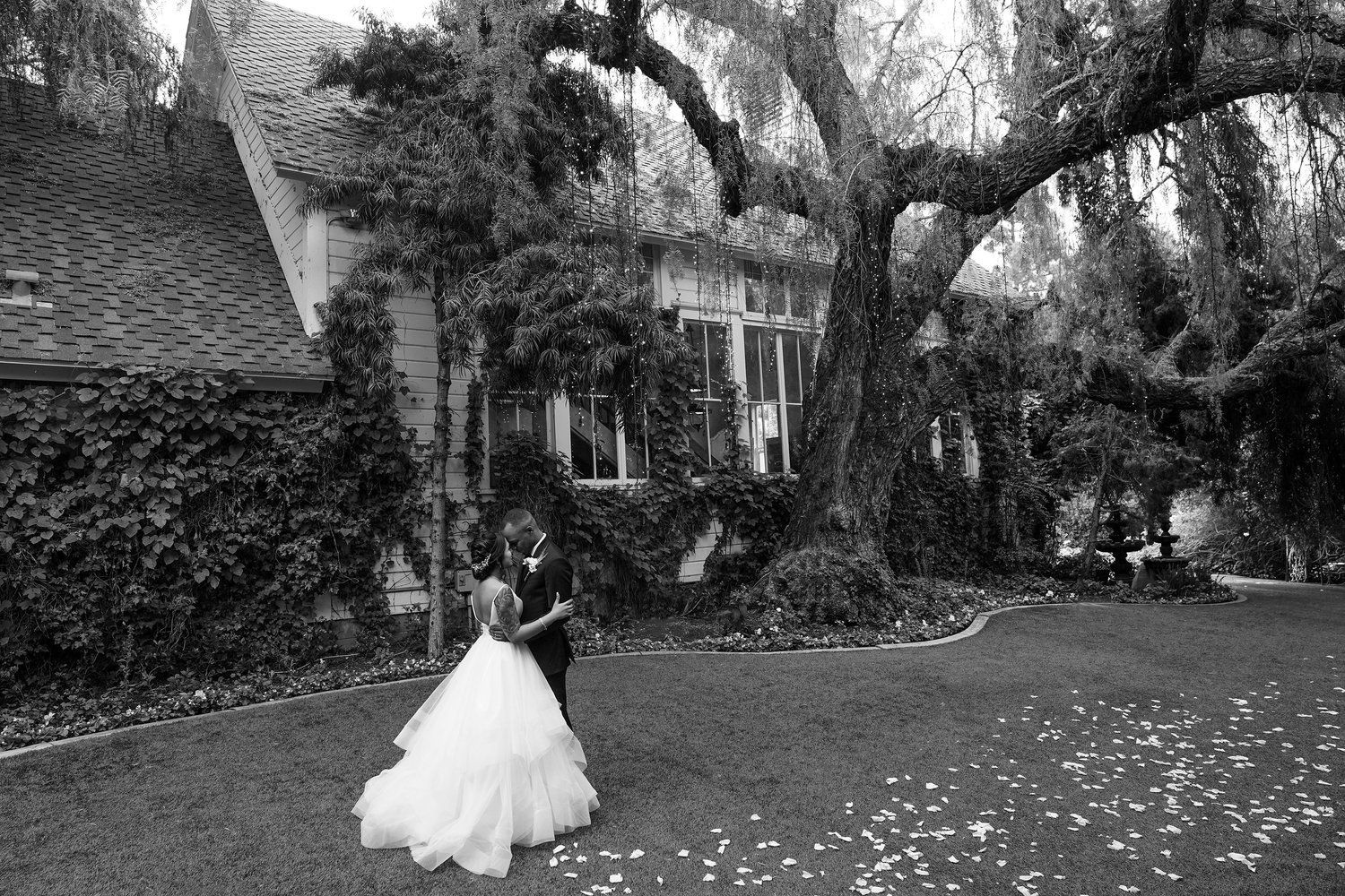 A black and white photo of a bride and groom dancing in front of a house.