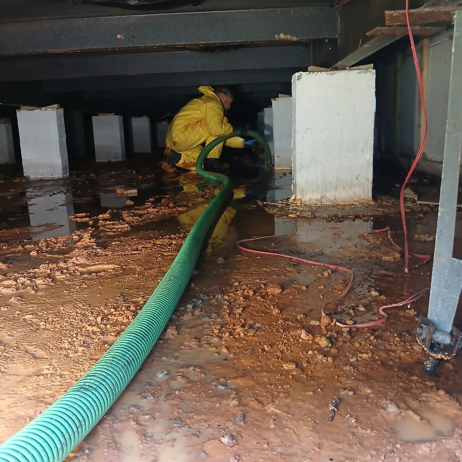 Person in yellow suit hoses muddy crawlspace under a building.