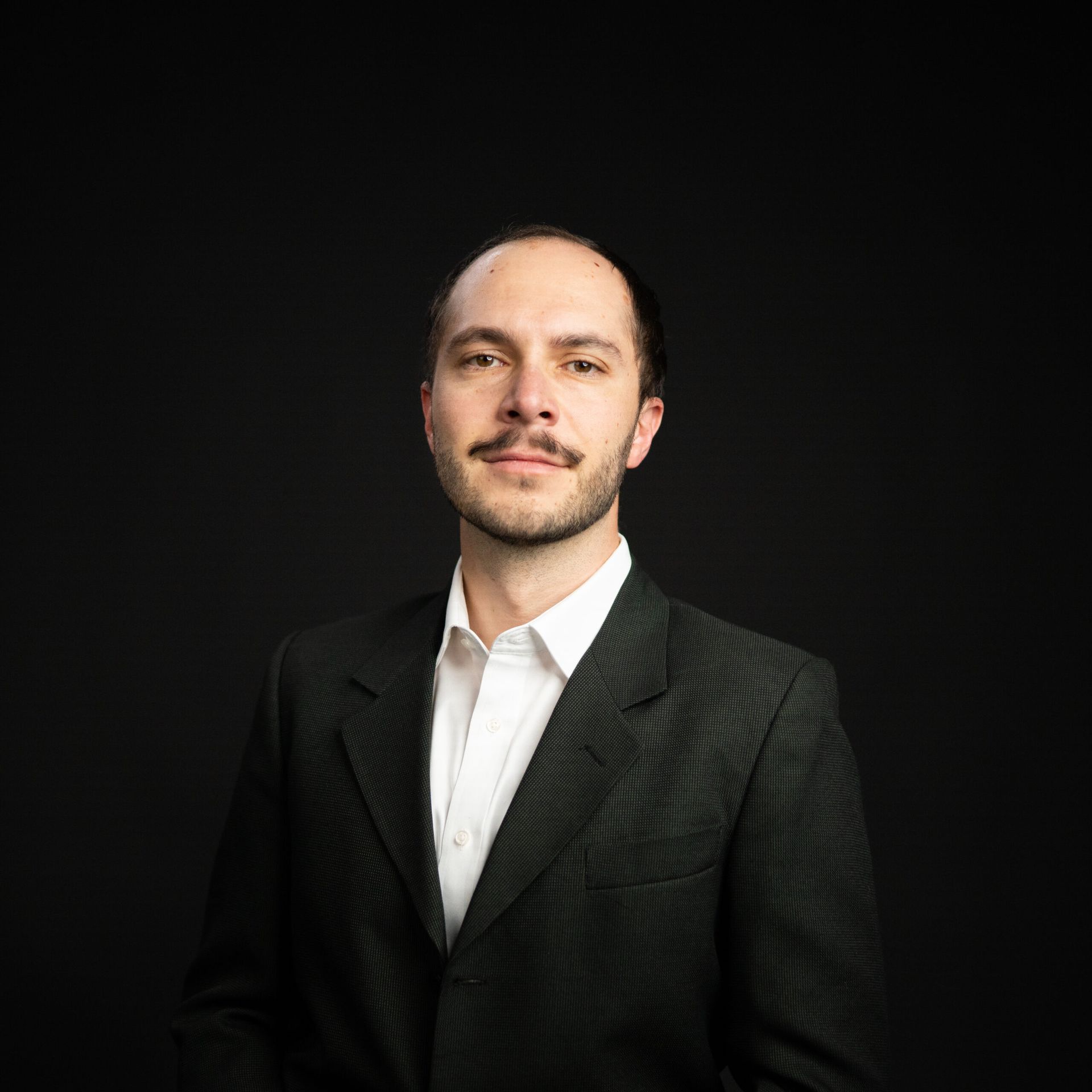 Man in a dark suit and white shirt, against a black background. He has a neutral expression and light facial hair.