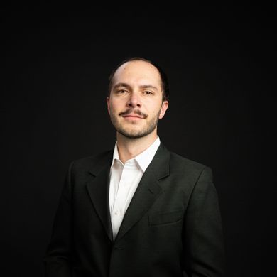 Man in a dark suit and white shirt, against a black background. He has a neutral expression and light facial hair.