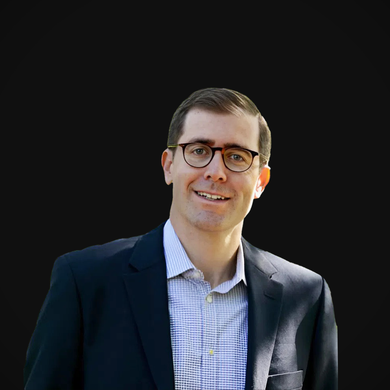 Man in glasses and suit jacket, smiling, with blue shirt, against black background.