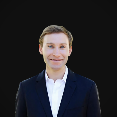 Man in navy blazer and white shirt smiles against a black background.