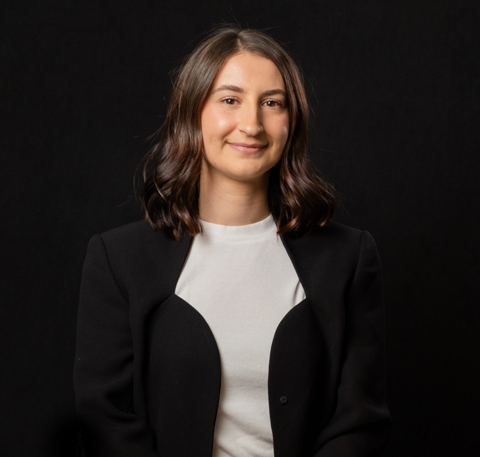 Woman with dark hair in a white shirt and black blazer smiles in front of a black backdrop.