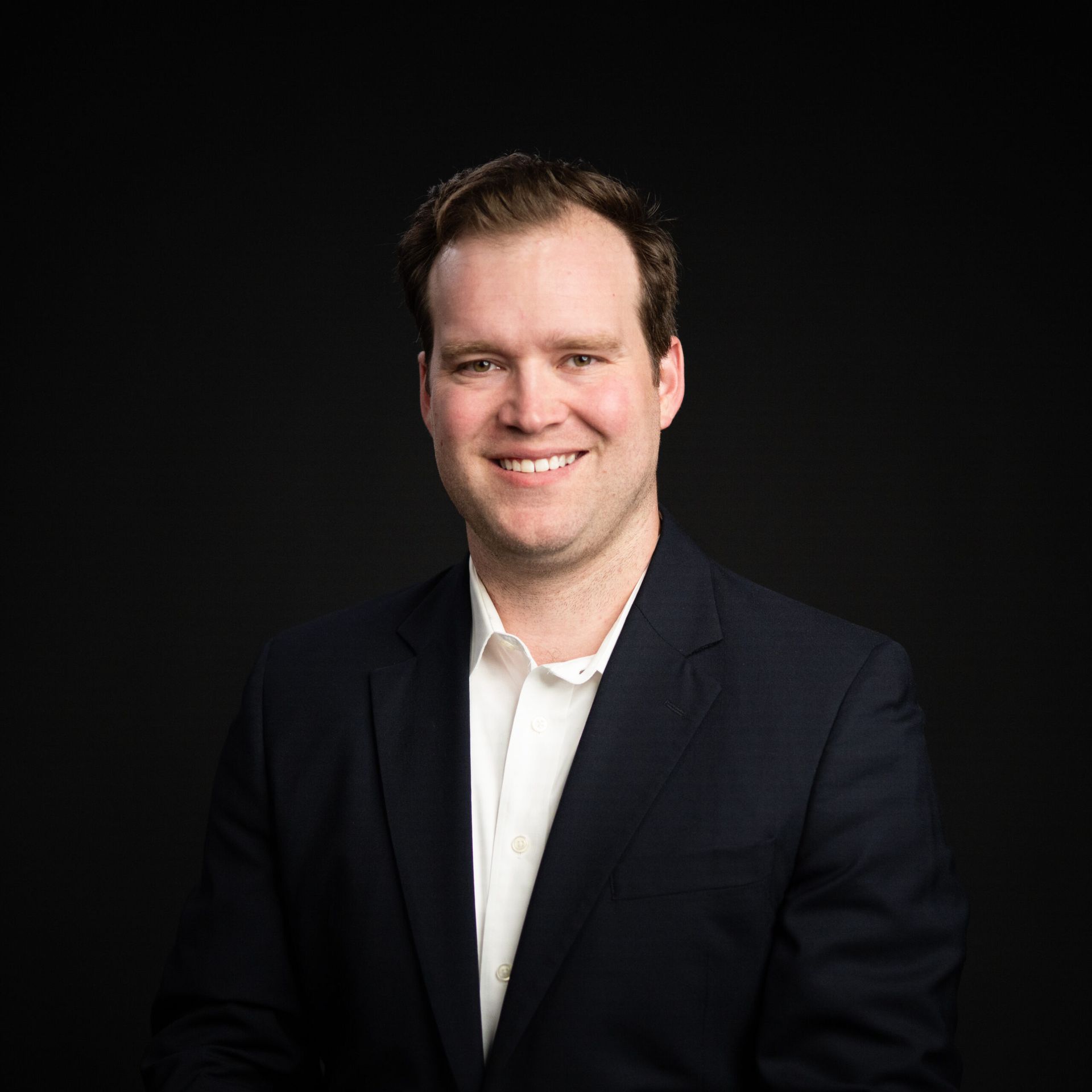 Man in a dark suit and white shirt, smiling, against a black background.