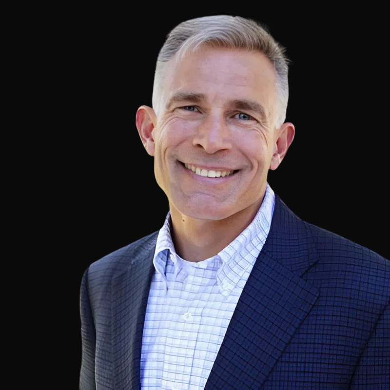 Man in blue blazer and patterned shirt smiling against a black background.