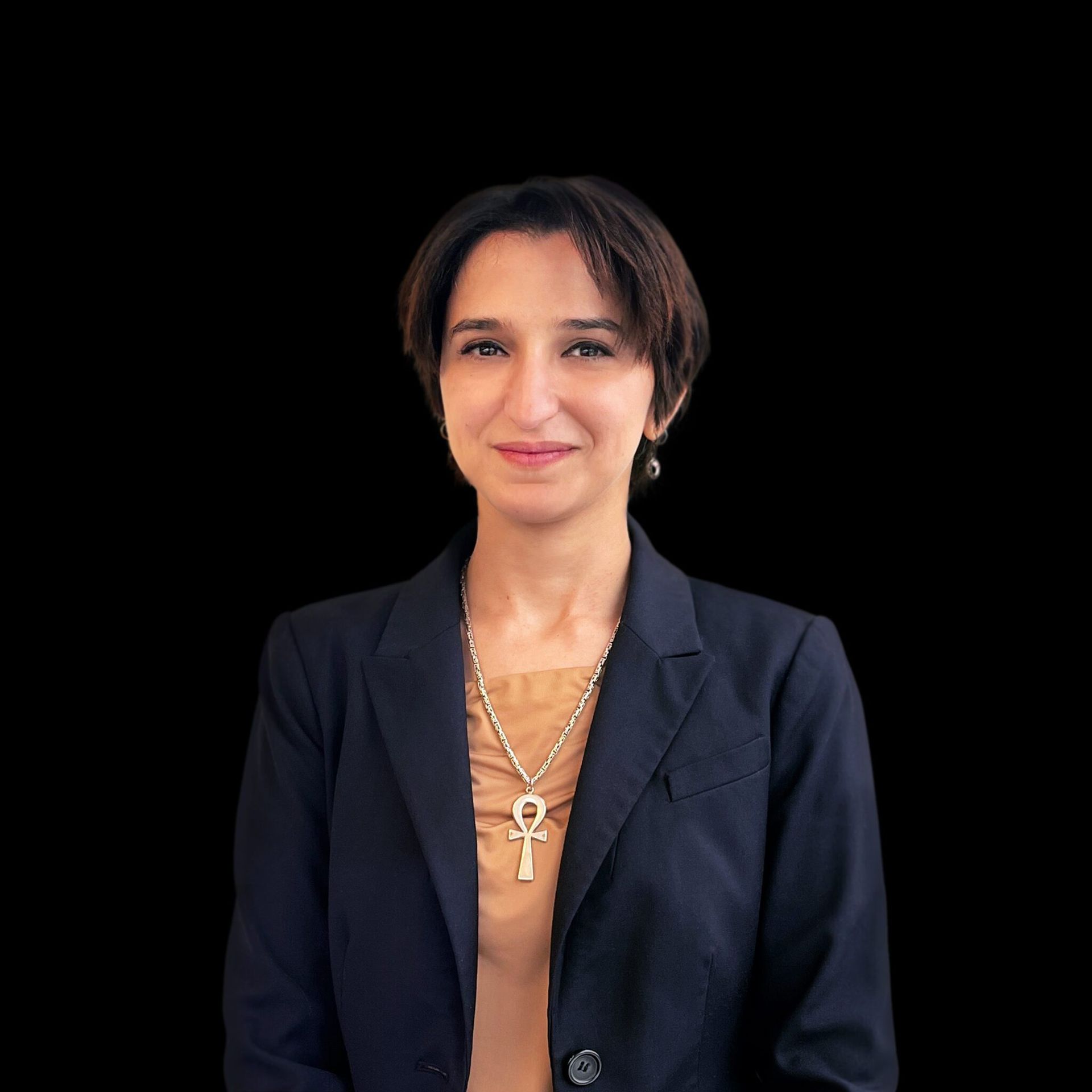 Woman with short dark hair, wearing a black blazer and necklace, smiling against a black background.
