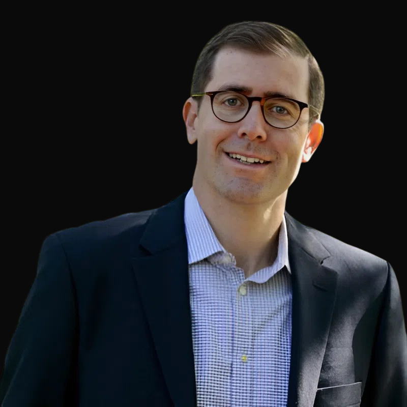 Man in glasses smiles, wearing a navy blazer and light blue patterned shirt against a black background.