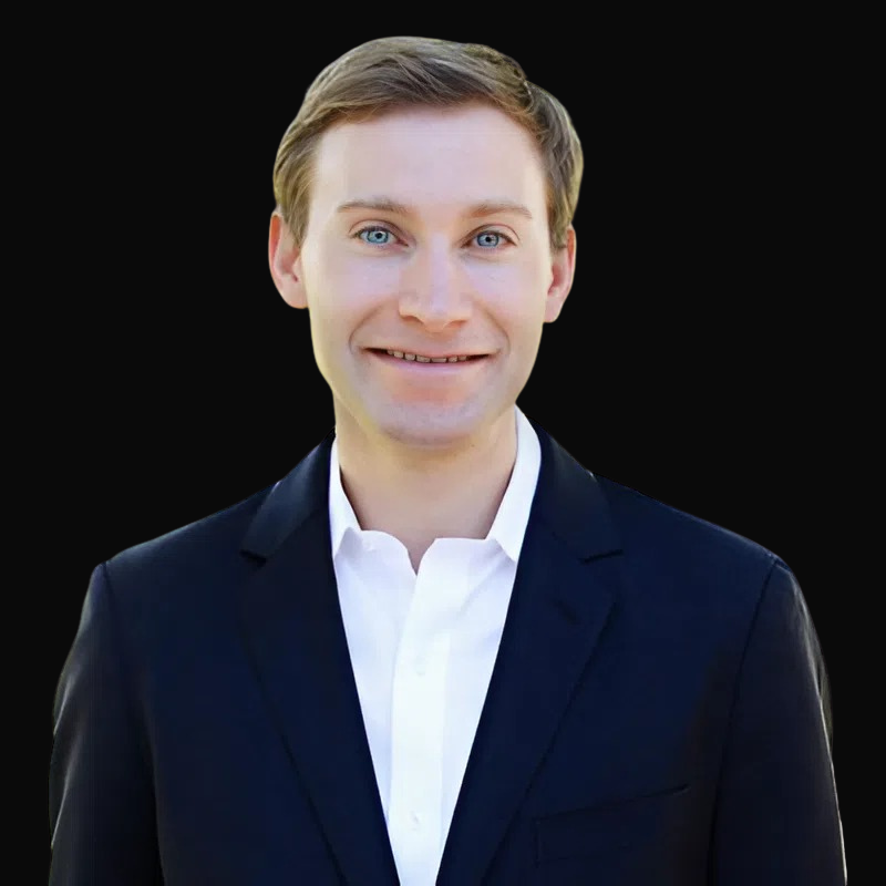 Man in a dark suit smiles, white shirt open at the collar, against a black background.