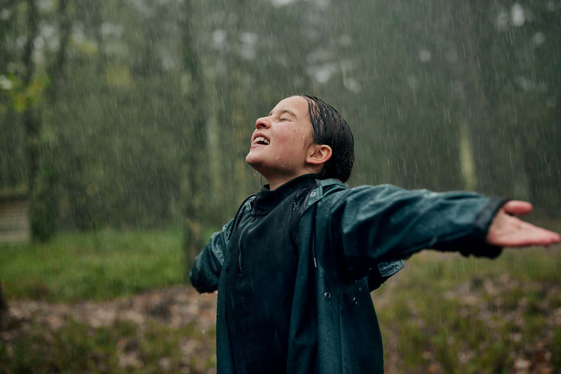 Een persoon in een donkerblauwe regenjas met uitgestrekte armen, lachend, genietend van de regen in een bos.